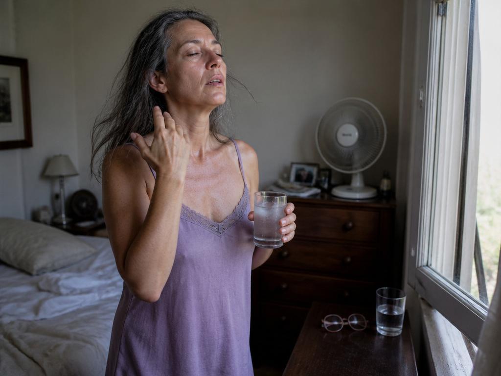 A woman in her early fifties by an open bedroom window fanning her flushed face during a hot flash, holding a glass of cool water
