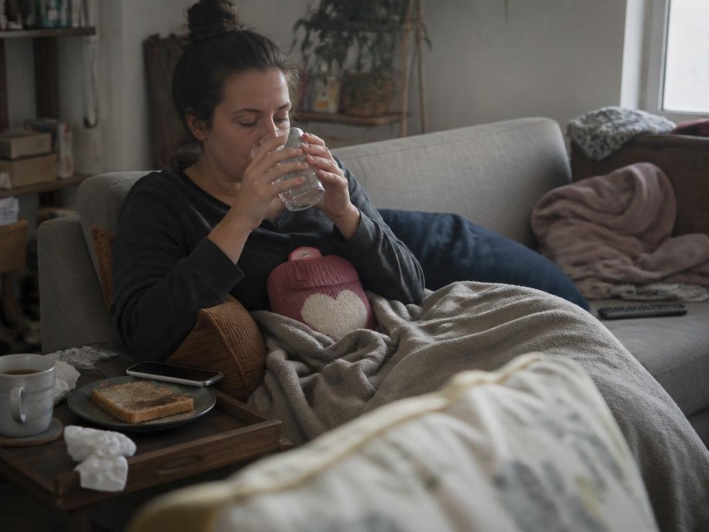A pale, tired woman curled under a blanket on a couch slowly sipping water with both hands, a hot water bottle against her stomach