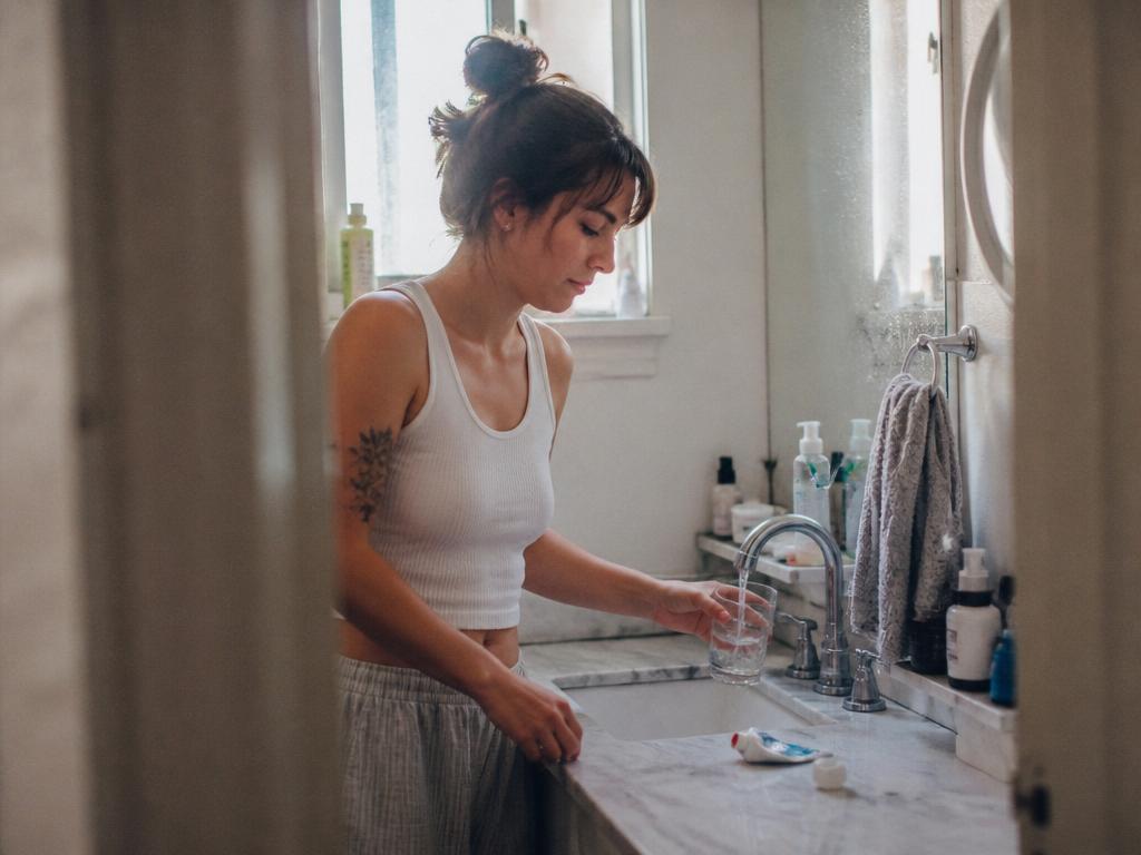 A woman filling a clear glass of water from the bathroom tap in bright daylight, toiletries crowding the counter and water spots on the mirror