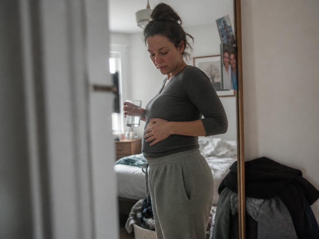 A woman standing sideways at a bedroom mirror resting a hand on her slightly bloated lower belly, a glass of water in her other hand